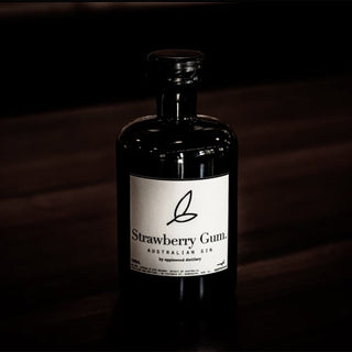 A dark glass bottle of Applewood Strawberry Gum Gin sits on a wooden surface. Moody lighting highlights its minimalist white label, featuring a simple leaf design. Crafted with native botanicals and wild berry essence.