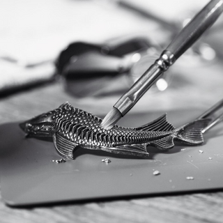 A close-up black and white photo shows a hand gently polishing the Beluga Celebration Limited Edition Vodka 700ml bottle, bringing out its metallic details, with a blurred background of tools and a work surface.
