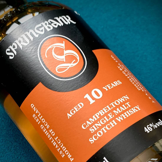 Close-up of a Springbank 10 Year Old Single Malt Scotch Whisky bottle, featuring its orange and black label with malt details and 46% ABV, displayed on a teal surface.