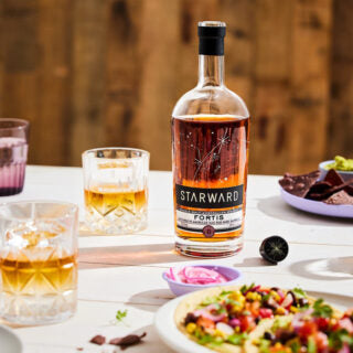 A bottle of Starward Fortis Australian Single Malt Whisky rests on a white table, surrounded by whisky glasses, a plate with salad-topped food, sliced red onions, chocolate ganache pieces, and set against a wooden background.