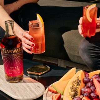 Two people with cocktails garnished with orange sit beside a bottle of Starward Nova Australian Single Malt Whisky on a table set with sliced melon, strawberries, grapes, and a platter evoking hints of vanilla and red berries.
