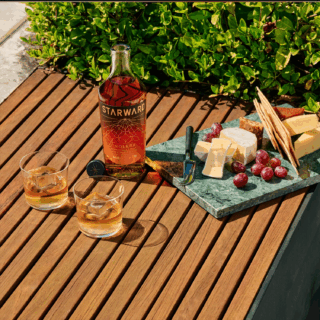 A bottle of Starward Solera Australian Whisky, two whisky-filled glasses, and a cheese board with ripe orchard fruits, grapes, crackers, and cheese are arranged on a wooden outdoor table amidst lush green foliage.