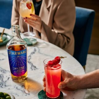 One person holds a bright pink cocktail with red berries; another holds a tropical-inspired drink. A bottle of Starward Two-Fold Australian Whisky rests on a round marble table.