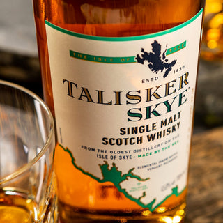 A close-up of the Talisker Skye Single Malt Scotch Whisky label, with a whisky glass and Isle of Skye map nearby. Warm hues and reflections evoke notes of citrus and coastal brine found in this single malt whisky.