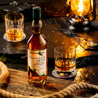 A bottle of Talisker Skye Single Malt Scotch Whisky sits on a dark stone with two filled glasses, a lantern, and thick rope nearby, all warmly lit—reflecting the whisky’s coastal brine and gentle peat smoke notes.