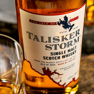 A close-up of a Talisker Storm Single Malt Scotch Whisky bottle with gold liquid visible next to a whisky glass. The label showcases its coastal origin from the Isle of Skye, Scotland.