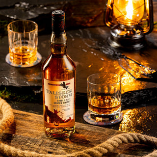A bottle of Talisker Storm Single Malt Scotch Whisky rests on a rustic surface next to two whisky glasses and a lantern, with coiled rope in the foreground and warm, ambient lighting.