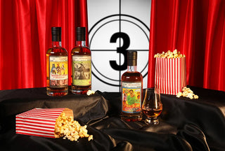 Three bottles of whiskey with a glass and popcorn on a black tablecloth against a red curtain backdrop.