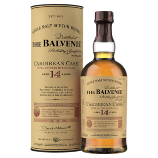 A bottle of The Balvenie Caribbean Cask 14 Year Old Single Malt Scotch Whisky stands next to its cylindrical box, both with tan and gold labels featuring black and red text that highlight the whisky's tropical toffee and vanilla notes.
