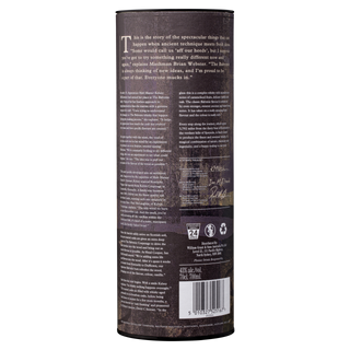 A tall, dark-textured cylindrical container for The Balvenie Stories The Sweet Toast of American Oak 12 Year Old Single Malt Scotch Whisky features product details, recycling symbols, a barcode, and white text near the base. Vanilla and oak spice notes are suggested.