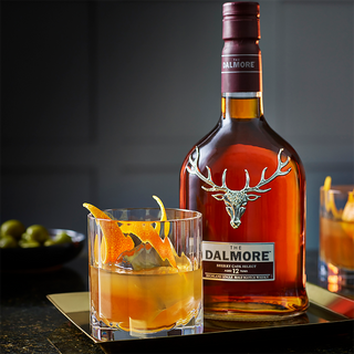 A bottle of The Dalmore 12 Year Old Sherry Cask Select Single Malt Scotch Whisky stands next to a glass with orange peel and caramelised oranges on a tray, set against a dark background.