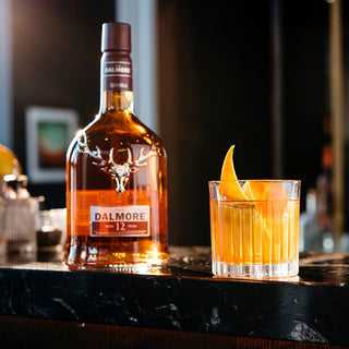 A bottle of The Dalmore 12 Year Old Single Malt Scotch Whisky sits on a dark bar beside a whisky cocktail garnished with orange peel, evoking spiced orange marmalade notes.