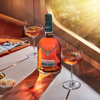 A bottle of The Dalmore 15 Year Old 2007 Vintage Single Malt Scotch Whisky rests on a train table with two whisky cocktails, snacks, and a clutch bag as sunlight streams in—orange marmalade and dark chocolate notes fill the air.