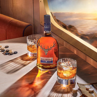 A bottle of The Dalmore 18 Year Old Single Malt Scotch Whisky sits on a wooden table with two glasses of whisky on the rocks, surrounded by game pieces and hints of spiced gingerbread, beside a window overlooking the ocean at sunset.