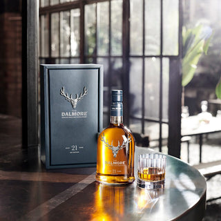 A bottle of The Dalmore 21 Year Old Single Malt Scotch Whisky sits with its box and a whisky glass on a sunlit table—large windows and gentle light highlight the scene, echoing subtle dark chocolate notes.