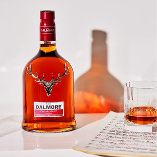 A bottle of The Dalmore Cigar Malt Reserve Single Malt Scotch Whisky sits beside a glass of single malt and a sheet of music, with a warm shadow falling across the white background.