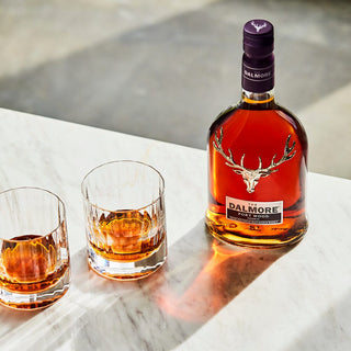 A bottle of The Dalmore Port Wood Reserve Single Malt Scotch Whisky rests on a marble surface beside two glasses, sunlight highlighting its amber hue and suggesting creamy caramel notes in each sip.