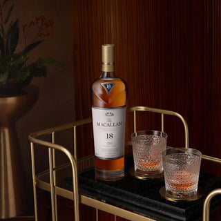 A bottle of The Macallan Double Cask 18 Year Old Single Malt Scotch Whisky rests on a gold bar cart beside two textured whisky glasses with ice, set against a backdrop of wooden panels and a potted plant for added style.
