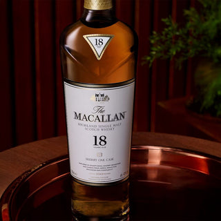 A bottle of The Macallan 18 Year Old Sherry Cask Single Malt Scotch Whisky (2024 Release) rests on a copper tray, with a blurred leafy plant behind it. The label highlights sherry cask aging, suggesting rich sherry sweetness and oak spice.