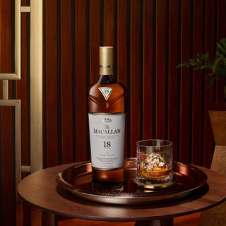A bottle of The Macallan 18 Year Old Sherry Cask Single Malt Scotch Whisky (2024 Release) and a glass with ice sit on a round wooden tray atop a table, set against dark wood paneling, evoking sherry sweetness and oak spice.