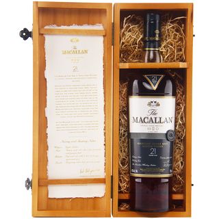 A bottle of The Macallan 21 Year Old Fine Oak Single Malt Whisky sits in an open wooden box with straw padding, accompanied by a paper describing its sherry notes and hints of vanilla.