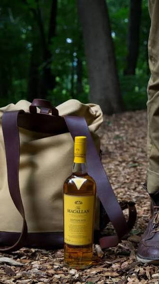 A bottle of Macallan Scotch whisky stands on a forest floor beside a tan canvas bag with dark brown straps, with a person's legs and boots visible in a wooded outdoor setting.