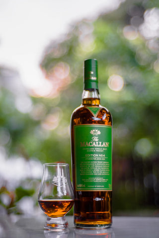 A bottle of The Macallan Edition No. 4 Scotch whisky stands on a reflective surface next to a small crystal whisky glass, with a blurred green outdoor bokeh background.