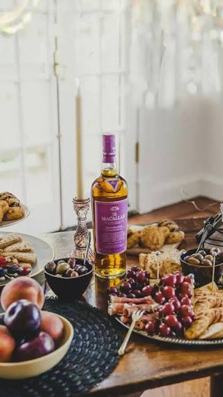 A bottle of Macallast whisky with a purple label stands on a wooden table surrounded by food, including grapes, olives, plums, and pastries, in a softly lit indoor setting.