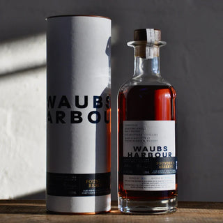 A bottle of Waubs Harbour Founders Reserve Tasmanian Single Malt Whisky with a cork top stands beside its white cylindrical packaging, both partly in shadow on a sunlit surface, against a light-colored wall.