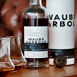A bottle of Waubs Harbour Founders Reserve Tasmanian Single Malt Whisky stands by its cylindrical packaging and cork, with two whisky-filled glasses evoking spiced pudding and salted pecan pie notes on a wooden surface.