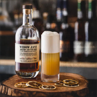 A bottle of Widow Jane 10 Year Old Bourbon Whiskey and a tall, foamy cocktail rest on a wooden slab beside dried citrus slices. Hints of charred oak and vanilla cream waft from the softly blurred bar shelf in the background.