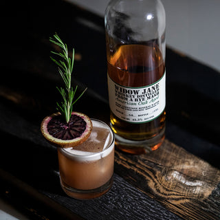 A glass of cocktail garnished with blood orange and rosemary sits beside a bottle of Widow Jane American Oak Aged Rye Whiskey on a dark wooden surface, evoking notes of spicy rye and mellow oak.