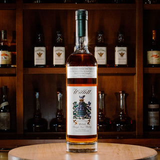 A bottle of Willett Family Estate 4 Year Old Rye Whiskey sits on a wooden surface, displaying notes of peppery spice. Behind it, shelves lined with various whiskey bottles appear blurred in the background.