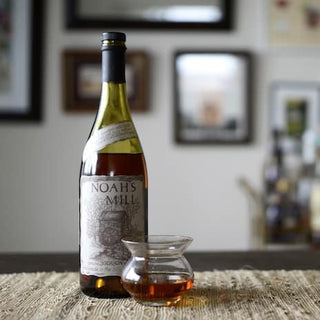 A bottle of Willett Family Estate Noah’s Mill Bourbon Whiskey sits next to a whiskey-filled glass on a woven placemat, exuding caramel and vanilla aromas, with blurred framed pictures visible in the background.