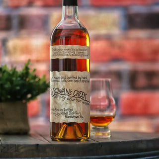 A bottle of Willett Family Estate Rowan’s Creek Small Batch Bourbon Whiskey with a handwritten-style label sits beside a whiskey glass on a table, accompanied by a small potted plant and set against a brick wall backdrop.