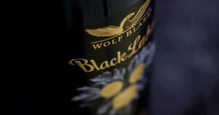 Close-up of a dark bottle label with gold lettering reading 'WOLF BLAD' and 'Black Label', plus a small white floral graphic, photographed against a dark blurred background.