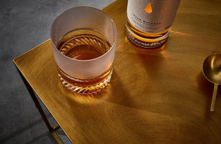 A glass of Writers Tears 2019 Cask Strength Irish Whiskey sits on a golden table next to its matching bottle and a gold-colored spoon, with toasted oak aromas swirling above the dark, textured surface.