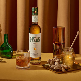 A bottle of Writers Tears Copper Pot Still Irish Whiskey sits on a yellow surface with a glass of whiskey, surrounded by creamy caramel notes, honey with a dipper, fresh ginger, green apples, and a brown curtain in the background.