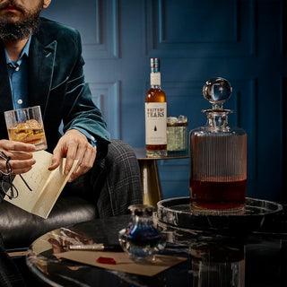 A person in a velvet jacket holds a glass of Writers Tears Double Oak Irish Whiskey, its vanilla caramel aromas filling the air, while sitting near a table with a decanter, envelope, and bottle against a dark blue paneled wall.