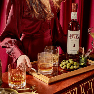 A woman in a burgundy dress holds a tray with Writers Tears Red Head Irish Whiskey, two glasses, fruit and nut chocolate, green olives, and cocktail picks on a wooden bar with a red curtain behind her.