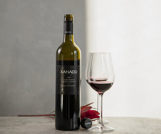 A bottle of Xanadu Cabernet Sauvignon red wine stands on a light stone surface beside a filled wine glass and a small red flower, against a soft gray and white background.