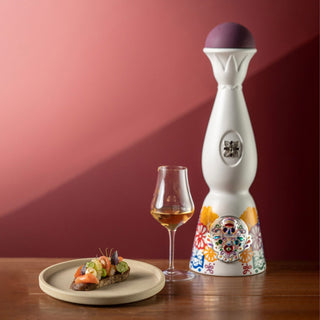 The Clase Azul Dia de Los Muertos 2022 Colores Limited Edition Tequila 1L, in a tall white bottle with colorful floral designs, sits beside a glass of amber tequila and gourmet food on wood, set against a red-pink gradient backdrop.