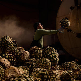 A worker in gloves lifts a large agave piña among harvested agave, with industrial equipment and dramatic lighting behind. Steam rises, evoking the artisanal process of crafting Don Fulano Imperial Extra-Añejo Tequila 700ml.