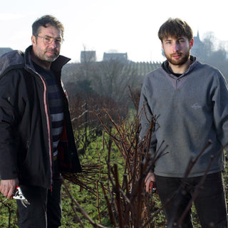 Two men stand in a vineyard holding pruning shears, ready to tend vines destined for Agrapart & Fils 7 Crus Extra-Brut NV—a fine sparkling wine with a crisp mineral finish. Bare grapevines, trees, and buildings are visible on the cloudy day behind them.