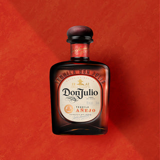 A bottle of Don Julio Anejo Tequila 700ml stands upright on a textured red background, its dark glass and cream label with gold and red accents evoking the rich caramel and agave notes inside.