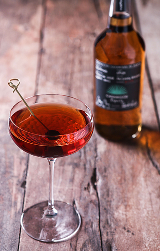 A coupe glass with a deep red cocktail and a dark cherry garnish sits on rustic wood, with notes of caramel and a blurred Casamigos Anejo Tequila 700ml bottle in the background.