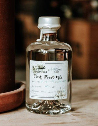 A clear glass bottle of Applewood x Catalina First Point Gin, infused with native botanicals, rests on a wooden surface indoors. The white wax seal and label show batch number, bottling date, strength, and volume details.