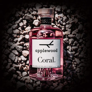 A 500ml bottle of Applewood Coral Gin, made with native botanicals and pink coral berries, features a cork stopper and white label, resting on small gray pebbles.