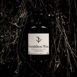 A bottle of Applewood Geraldton Wax Gin 500ml rests on dry grass and purple flowers, its white label centered—showcasing native botanicals and delicate floral citrus notes.