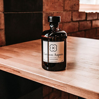 A bottle of Applewood Lemon Aspen Gin 500ml rests on a wooden countertop in a warmly lit room with brick and dark walls.